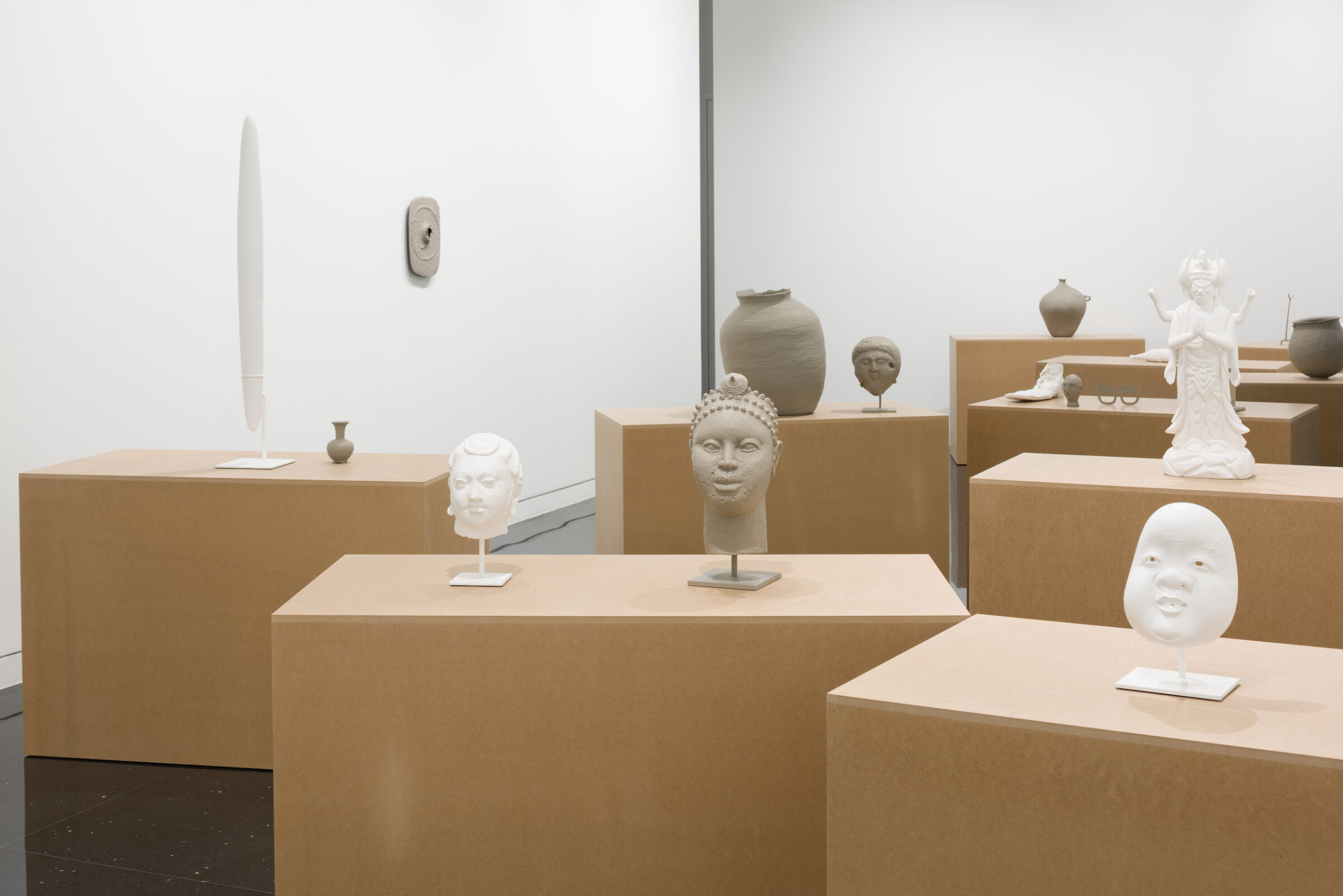 Ten pedestals resembling cardboard boxes in a white gallery space. Atop the closest pedestal are two unglazed ceramic sculptures of a mask and a head. The mask to the left of the head appears to be fired while the head appears unfired. Behind these sculptures, an unglazed and unfired jug sits on another pedestal. Several more ceramic objects appear scattered on the tops of each pedestal in the gallery.