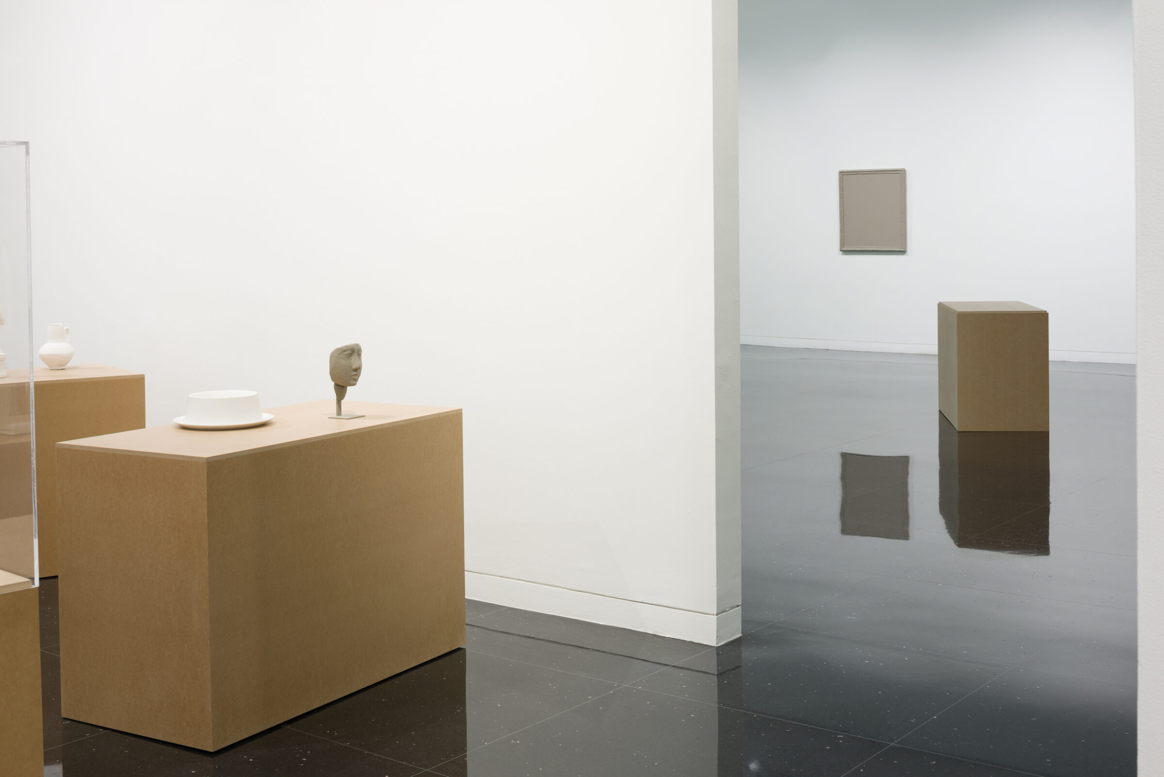 A pedestal resembling a cardboard box sits next to a white, freestanding gallery wall. Atop the box is an unfired clay mask, to the left of which sits a fired but unglazed ceramic hat. To the right and on the other side of the freestanding wall, another pedestal is visible next to a gray square installed on the gallery wall.