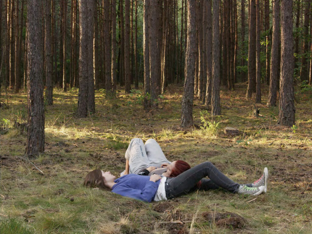 Two girls lay in the middle of a forest, one resting their head on the others thigh.