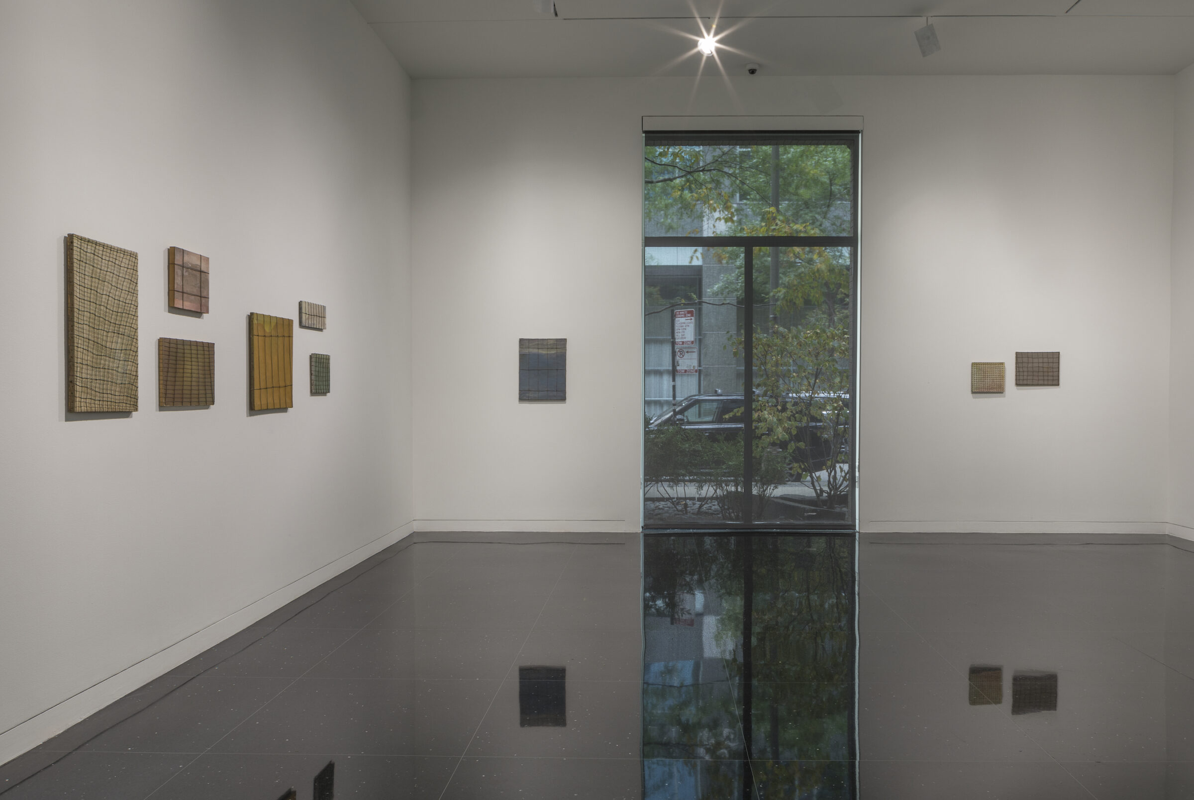 Installation view of a white walled gallery with two dimensional works of varying sizes arranged on the walls. Most works appear to have a wire grid laid over the canvas.