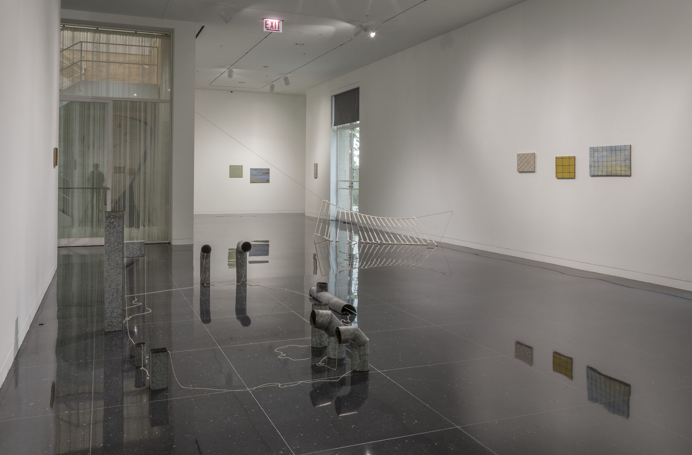 Installation view of a white-walled gallery with sculptures suspended from the walls and on the gallery floor. A series of bent exhaust pipes arranged in a circle are visible.