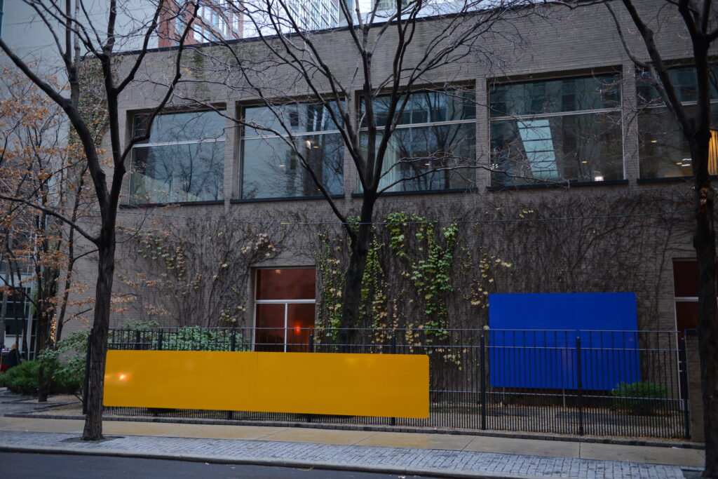 A long yellow rectangle hangs on a fence in front of a two-story brick building while a smaller blue rectangle covers part of the ivy-covered wall.