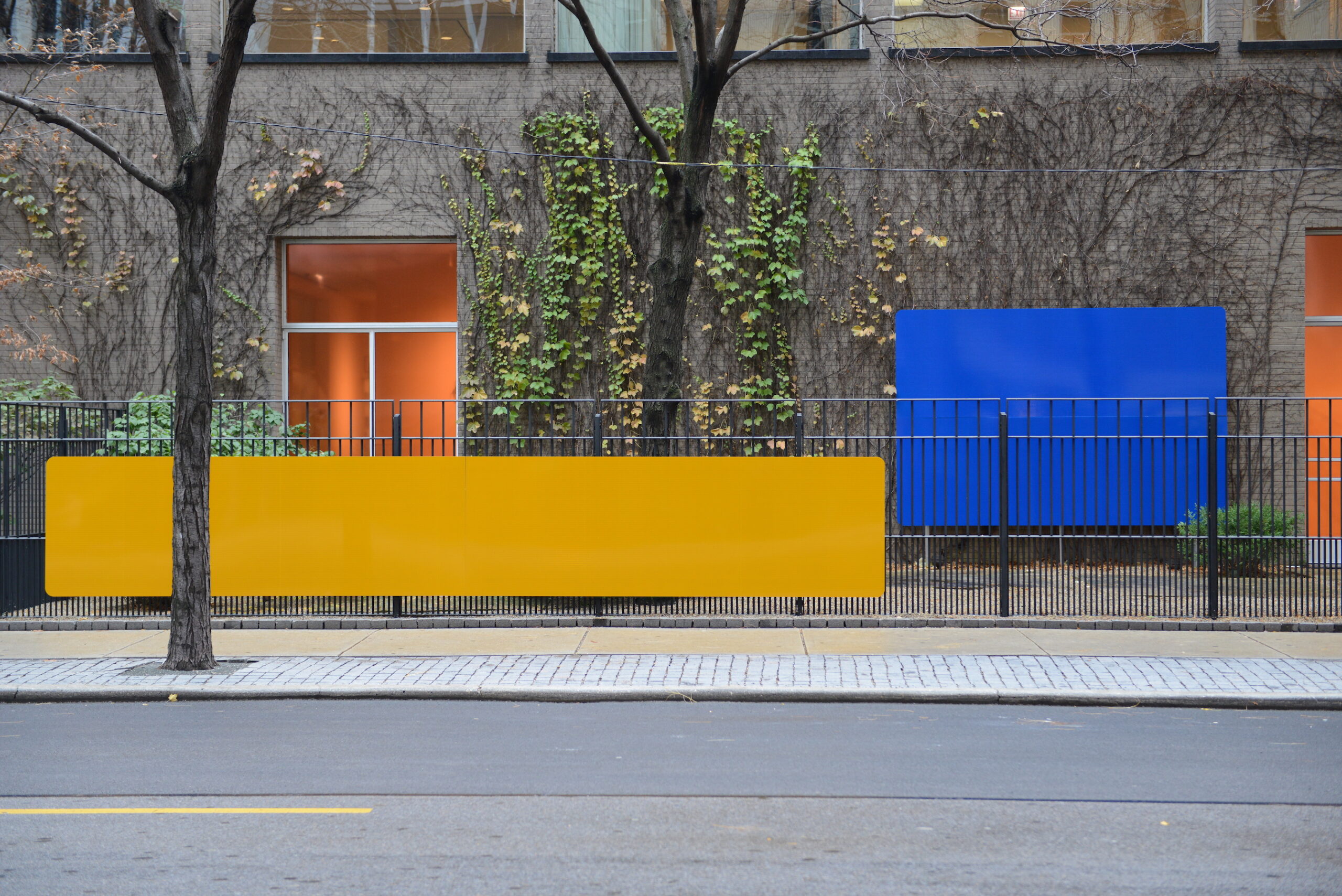 A long yellow rectangle hangs on a fence in front of a brick building while a smaller blue rectangle covers part of the building's ivy-covered wall.
