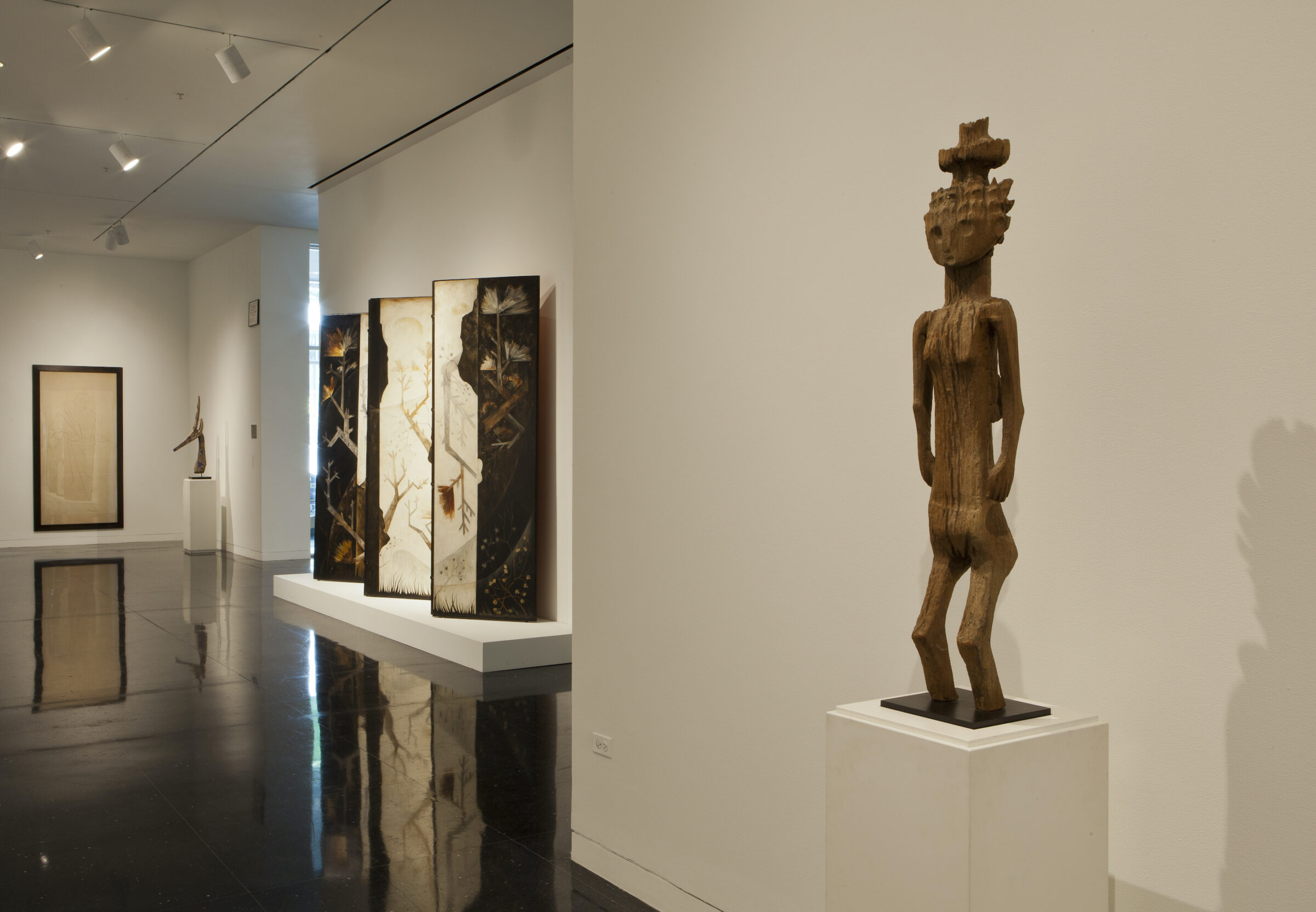 A soft lit gallery featuring a wooden carved sculpture of a female figure balancing an object on her head and standing on a square white pedestal.
