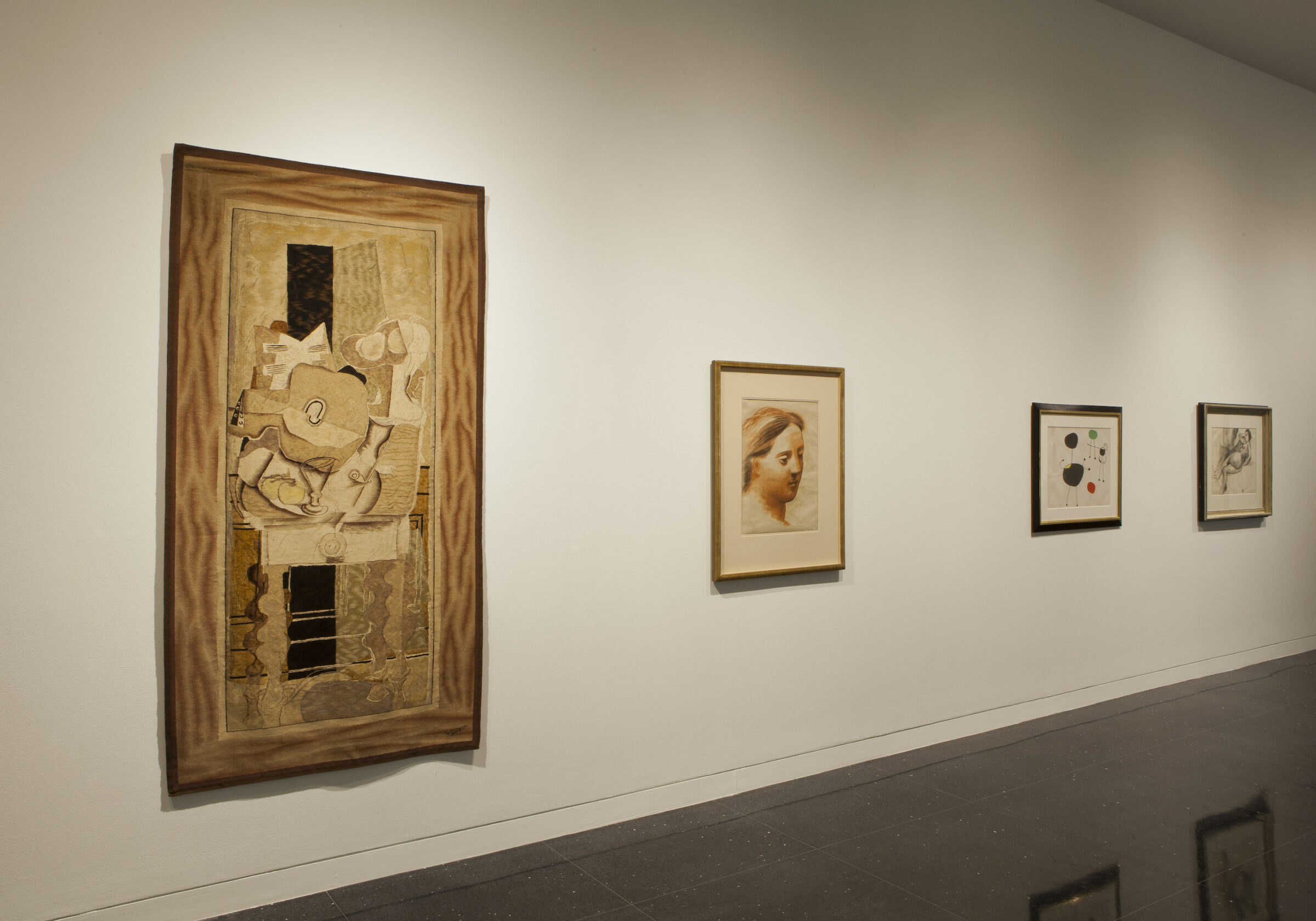 Soft lit, white walled gallery featuring a brown and beige tapestry reproduction of a Braque still life, a picasso drawing of a woman's head and neck, and an abstract picture by Joan Miro, hanging in a horizontal line.