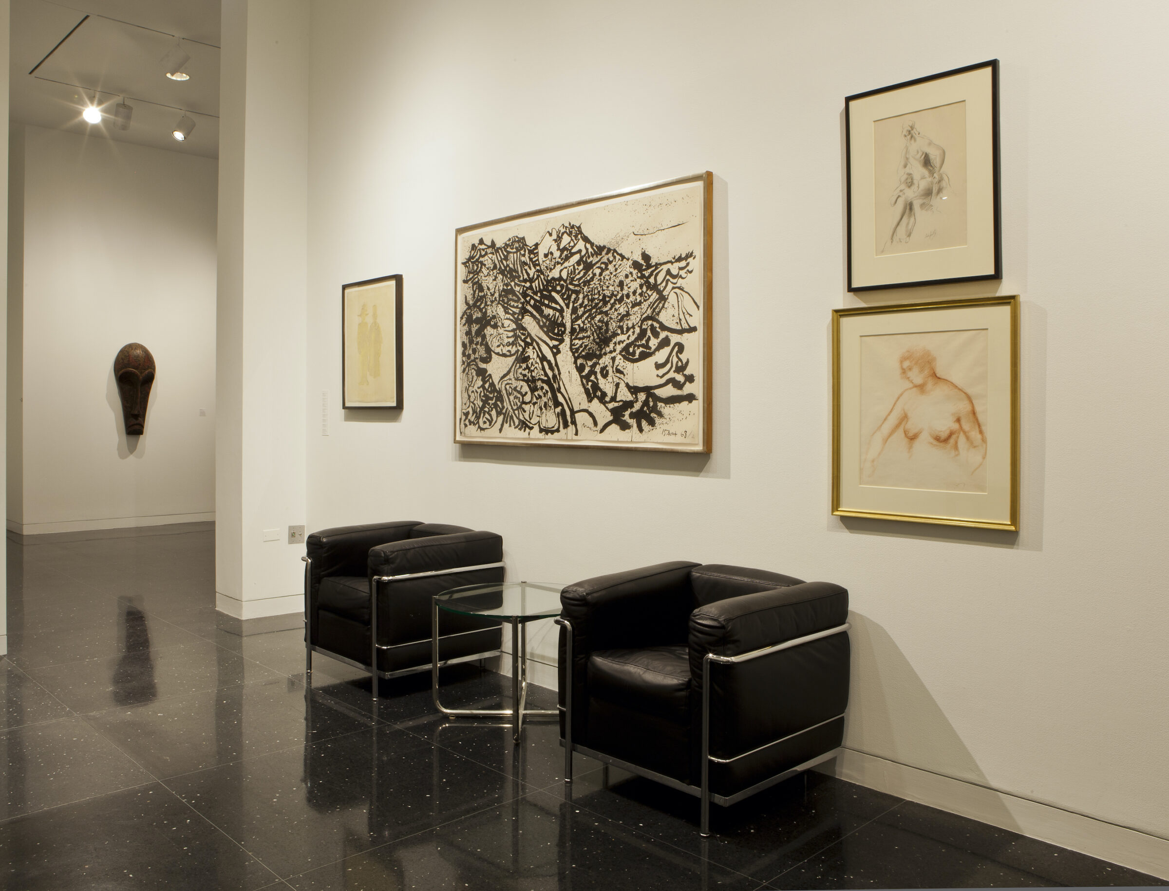 Two square-shaped cushioned arm chairs separated by a glass top table sit against a white gallery wall. Hanging on the wall between the two chairs is an abstract painting of black forms by Pierre Alechinsky. On either side of the painting are a series of framed sketches.
