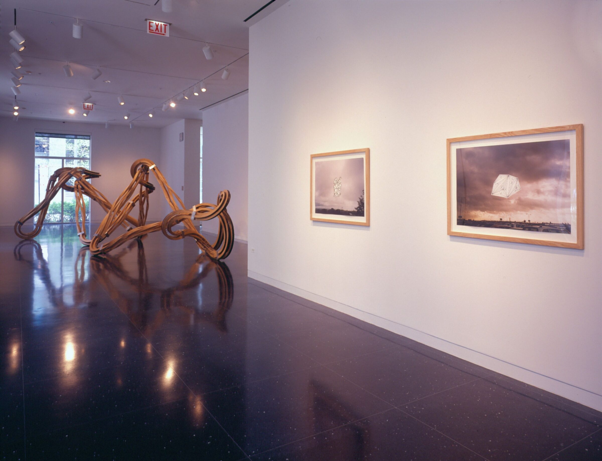 A white gallery space with two framed black and white photographs on a wall next to the viewer and a large bronze sculpture occupying the majority of the distant gallery space.