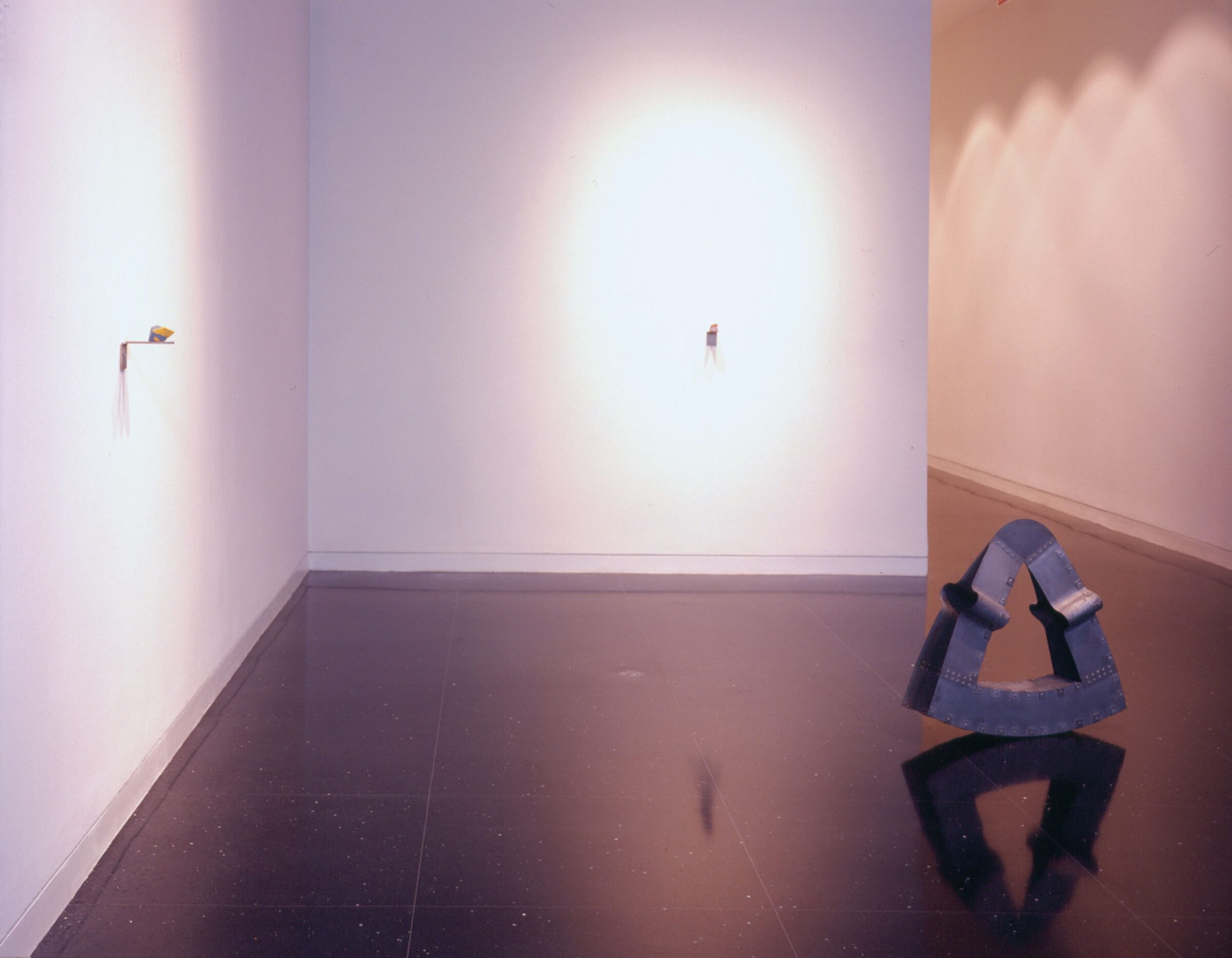 A white gallery space with a steel rocking sculpture on the floor and two very small rocklike sculptures installed on the walls.