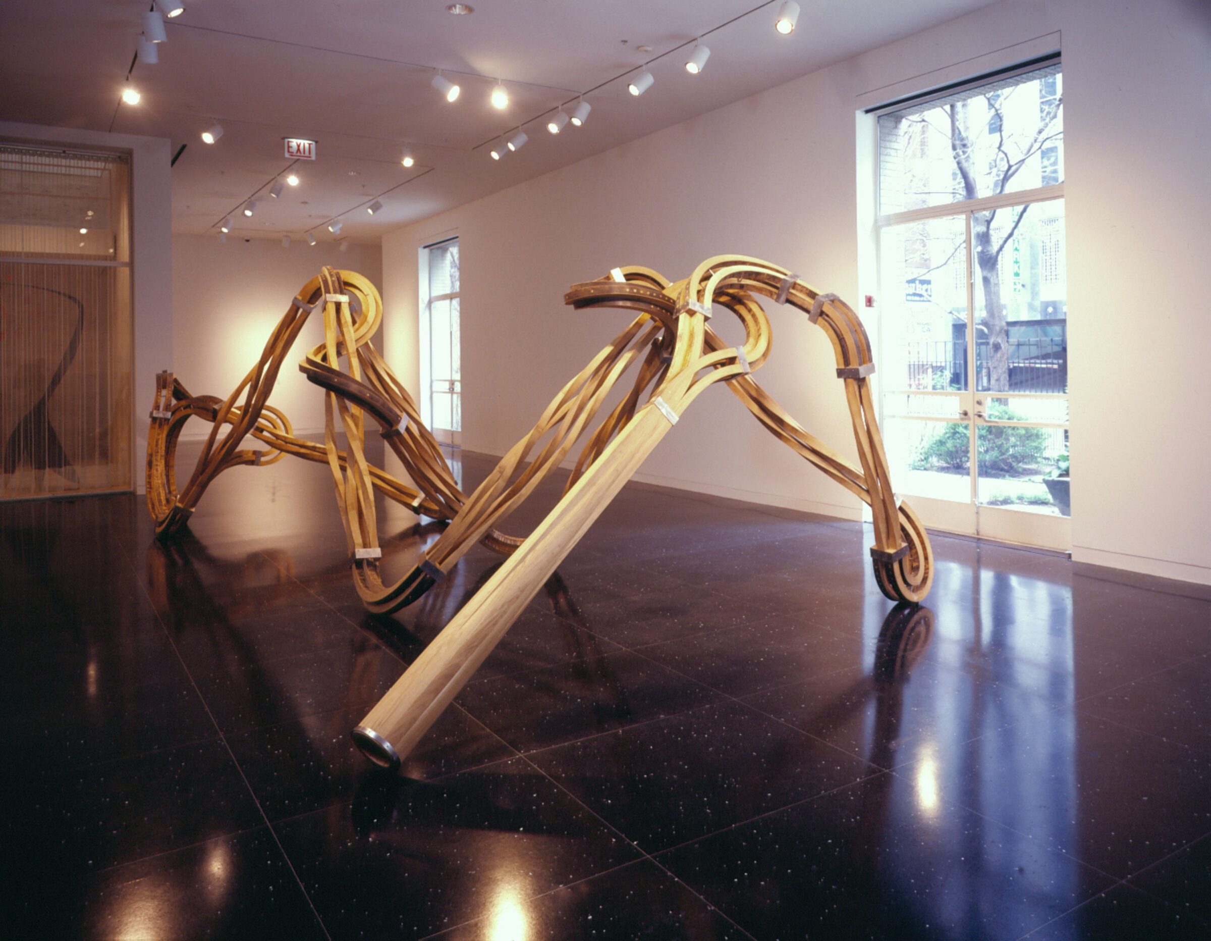 A large, tendril-like bronze sculpture occupies the majority of a white gallery space.