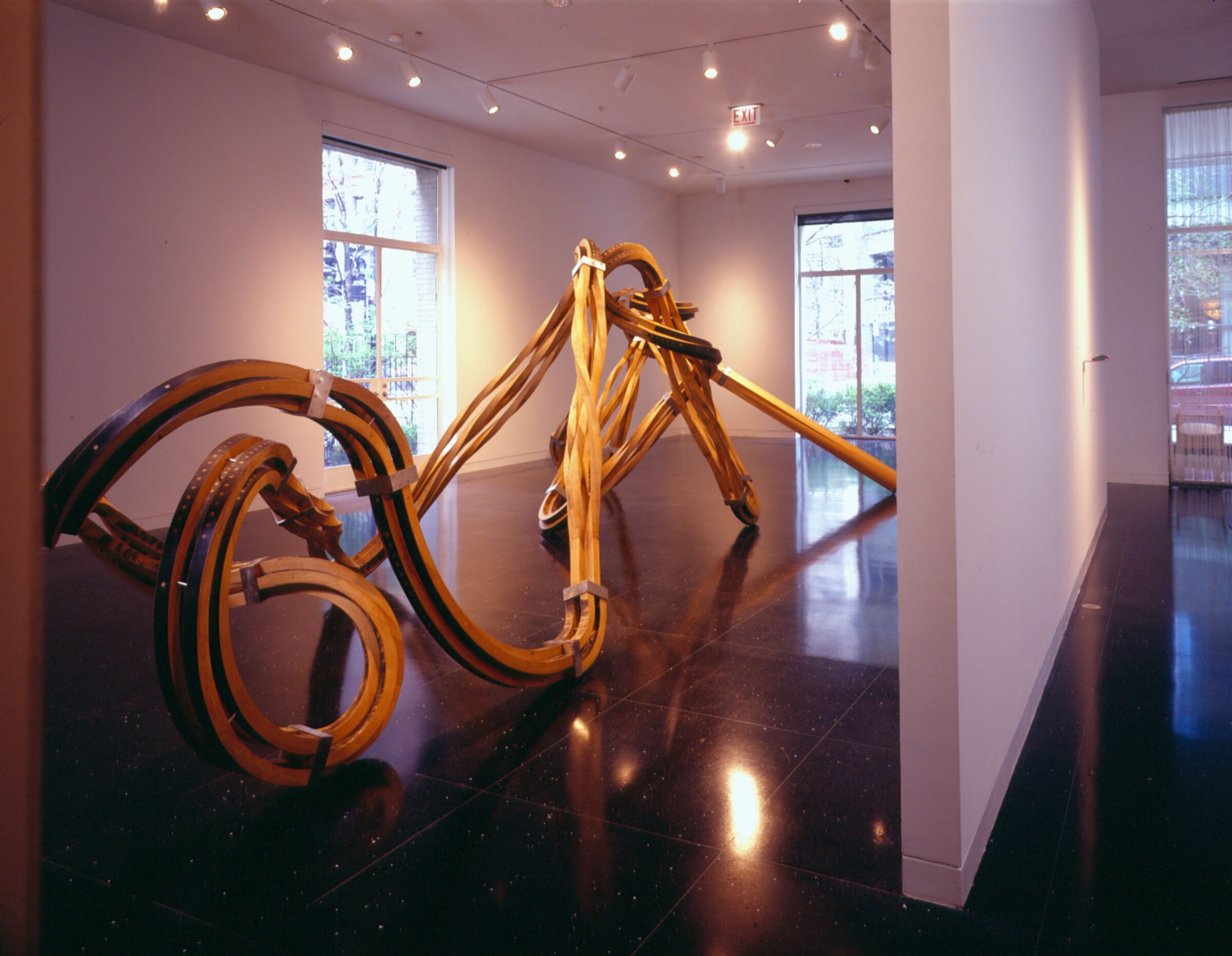 A large, tendril-like bronze sculpture occupies the majority of a white gallery space next to a freestanding wall.