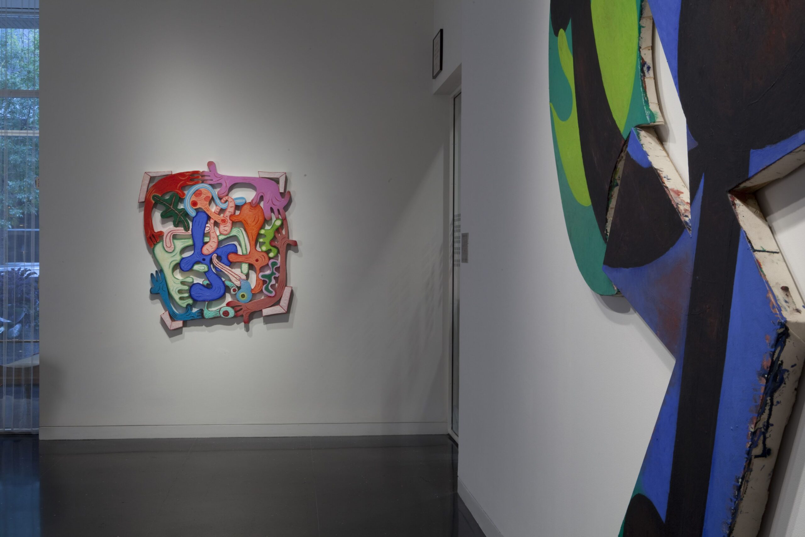 A white gallery space with two large, abstract artworks in colorful prints cut and carved into amorphous shapes. In the artwork furthest from the viewer, several colorful arms interlock to form a border or frame around the work.