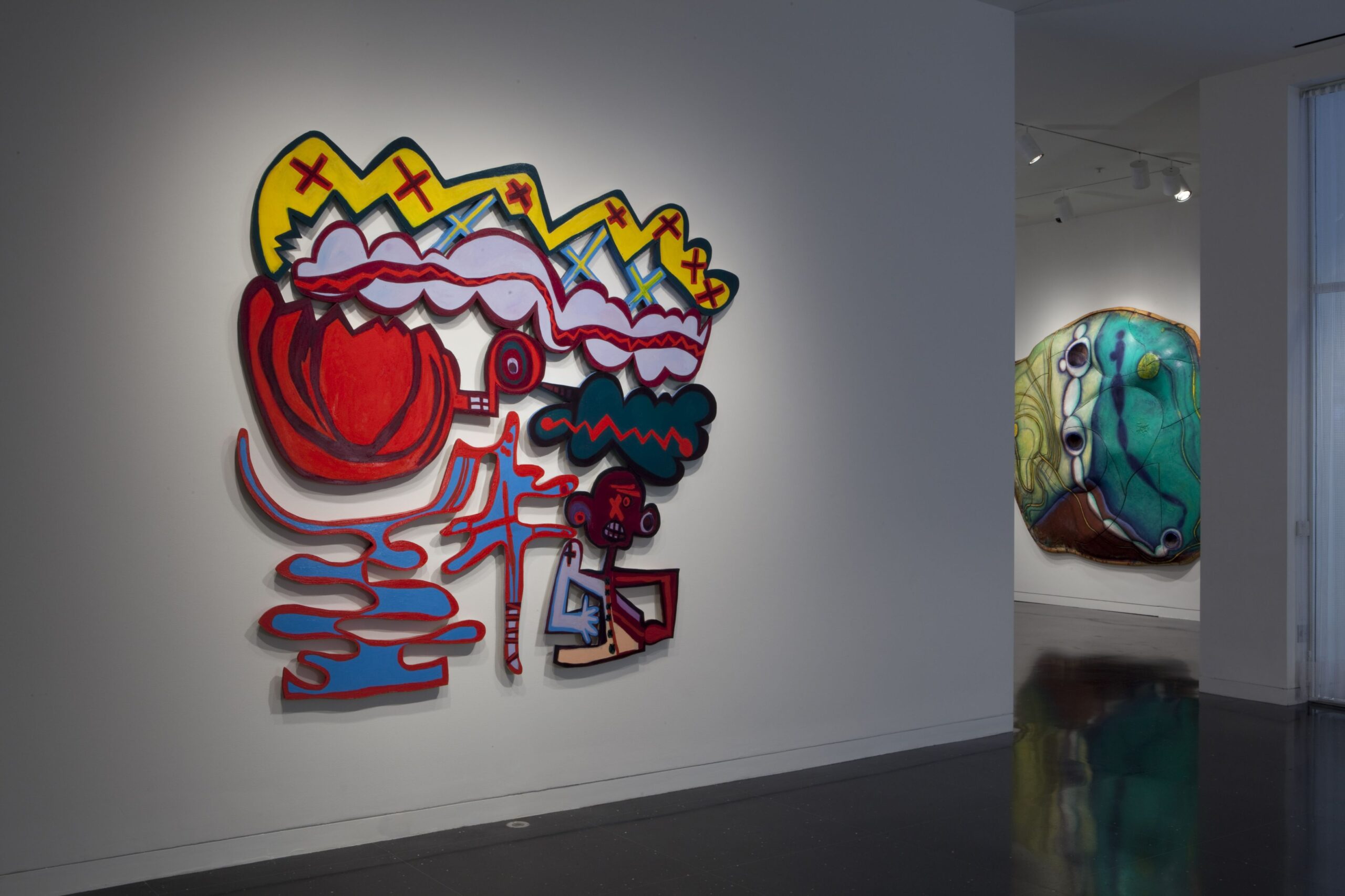 A white gallery space with two large, abstract artworks in colorful prints cut and carved into amorphous shapes. In the closest artwork to the viewer, a stylized humanoid figure is visible.