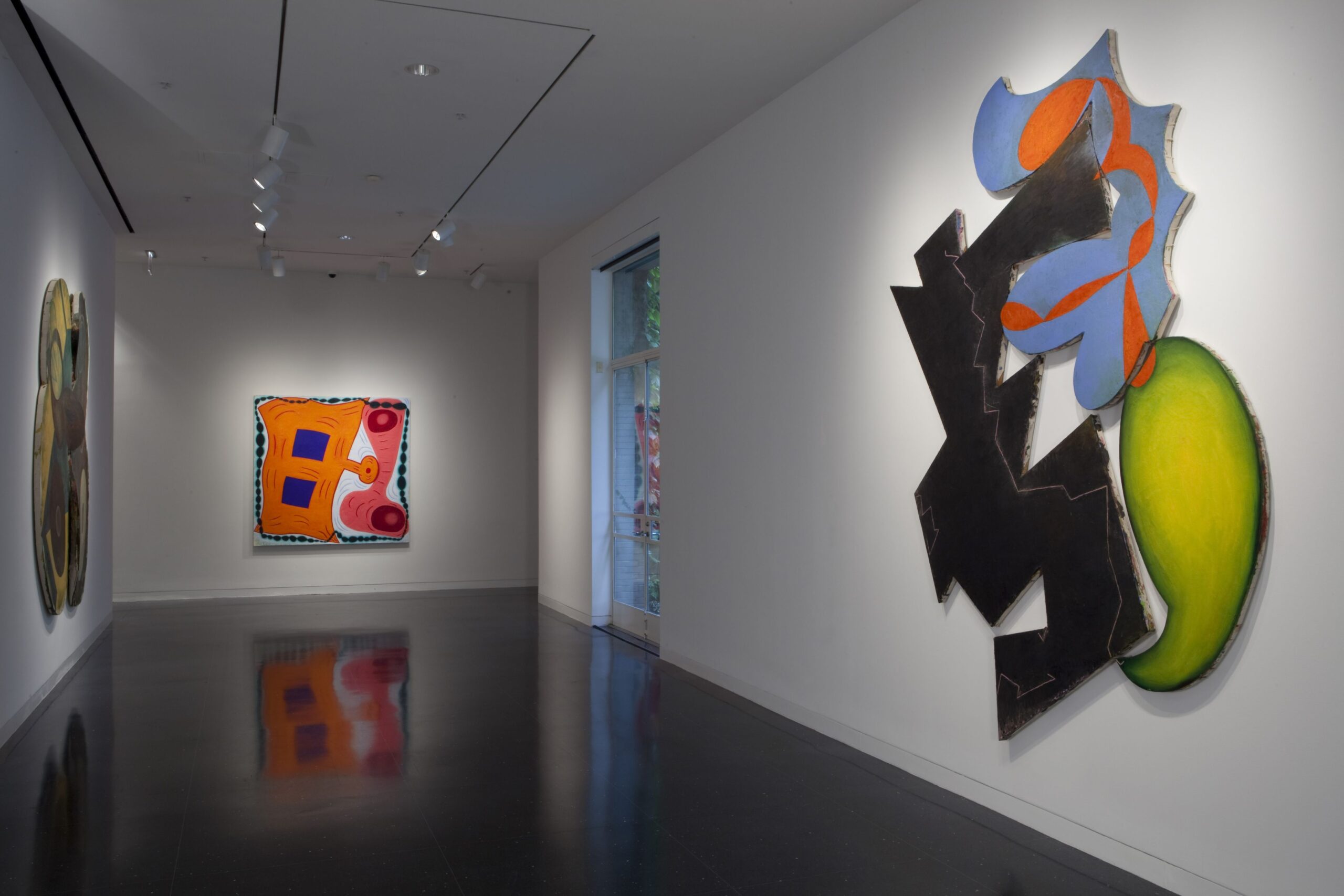 A white gallery space with two large, abstract artworks in colorful prints cut and carved into amorphous shapes. On a far wall is a large, square, mostly orange abstract painting.