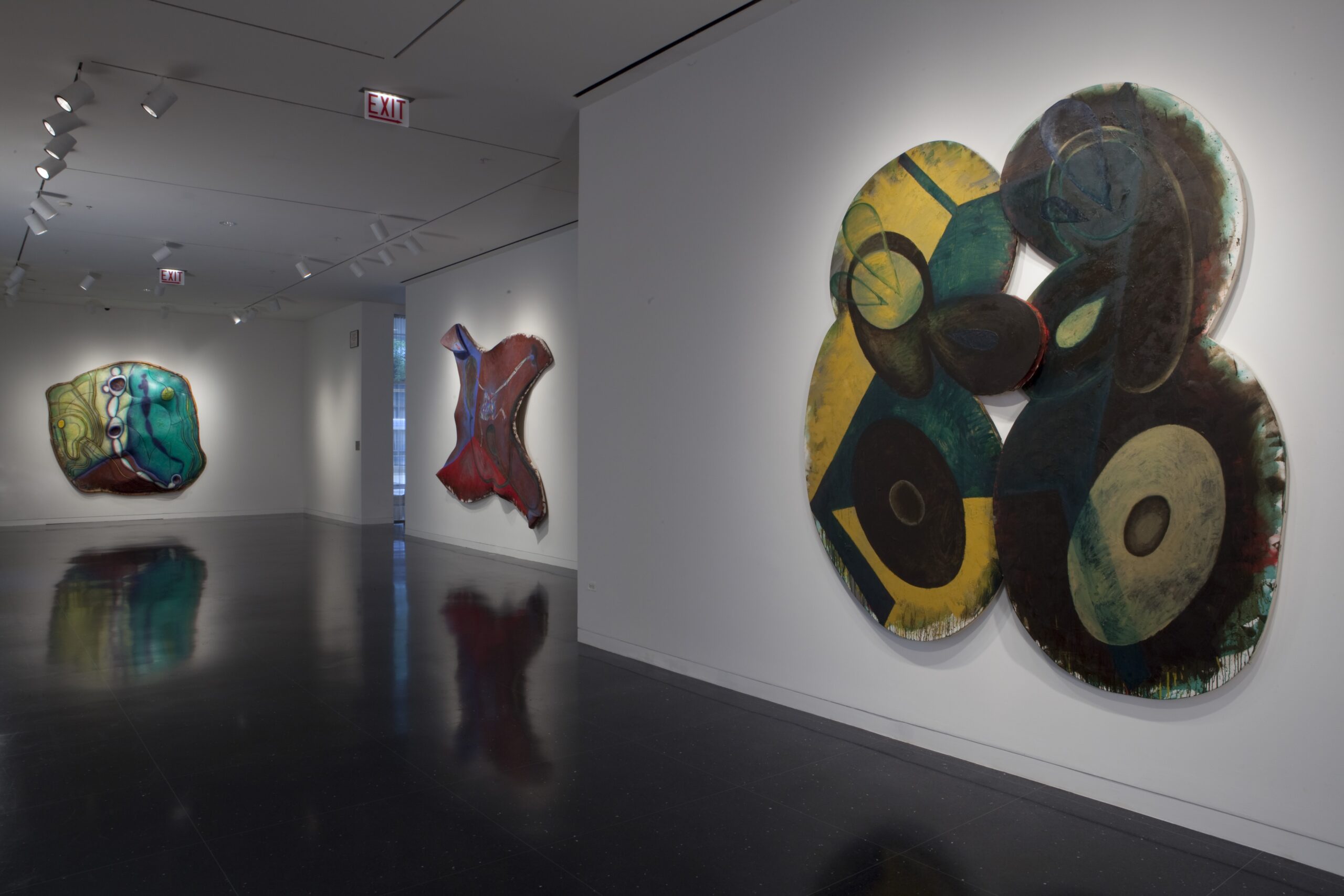 A white gallery space with three large, abstract artworks in colorful prints cut and carved into amorphous shapes.
