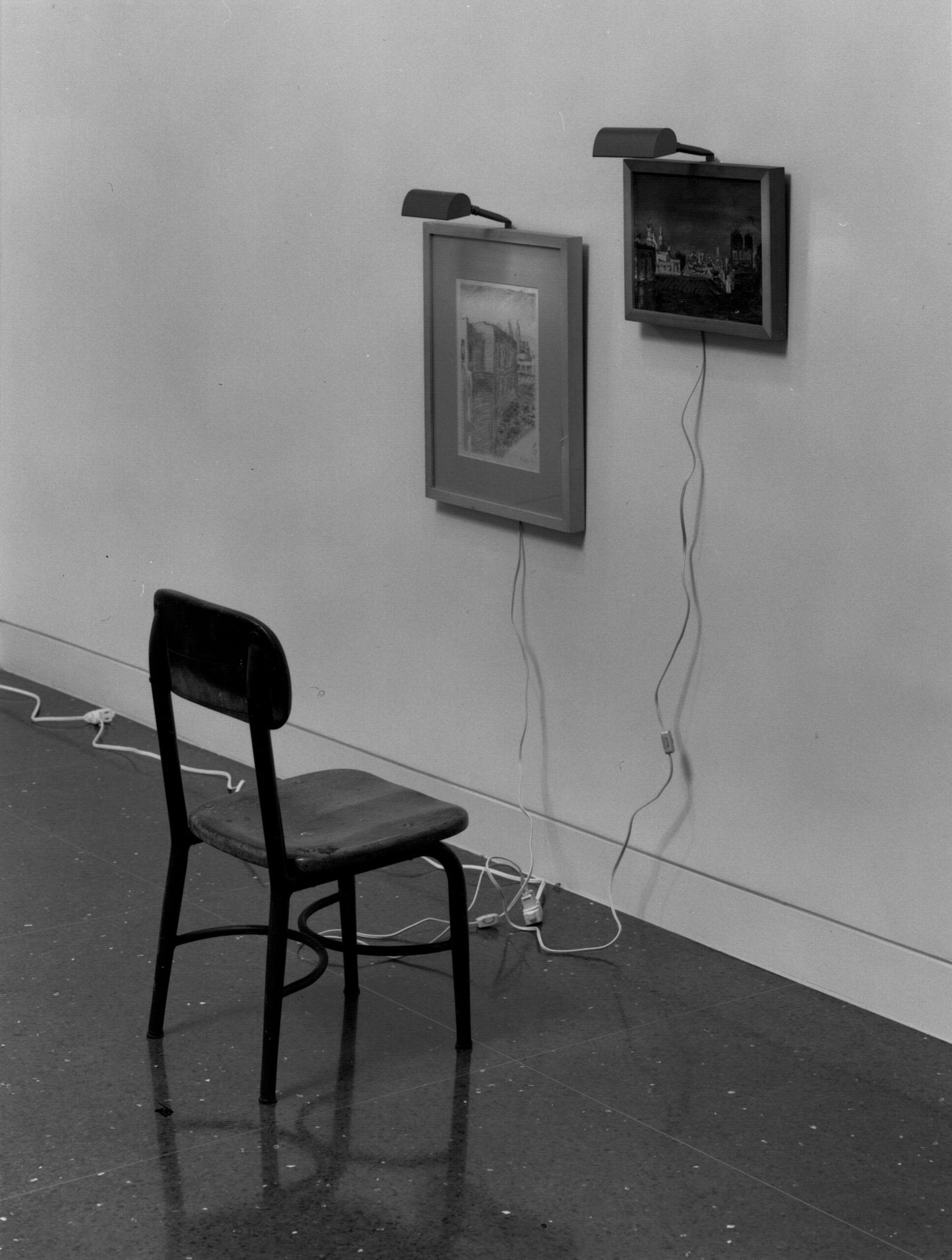 A white gallery wall on which two small framed images are installed with study lights right above them. Power chords run from the bottom of each piece, and a small black chair is placed in front of the images.