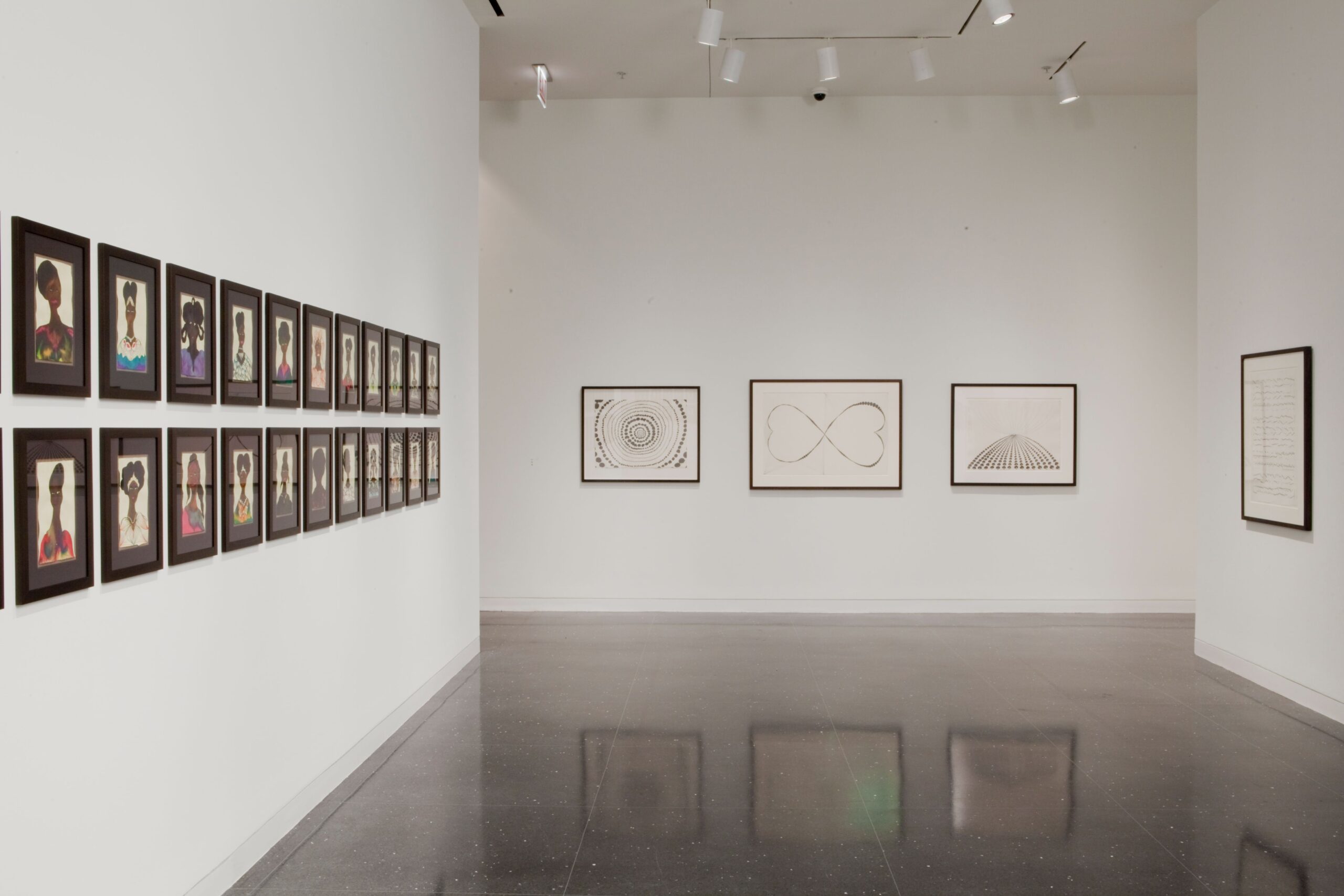 Three white gallery walls on which framed artworks hang. From left to right, the viewer can see Two rows of twelve small watercolor portraits of Black subjects in black frames, three framed and somewhat abstract drawings on white paper, and one framed drawing resembling writing or cartography.