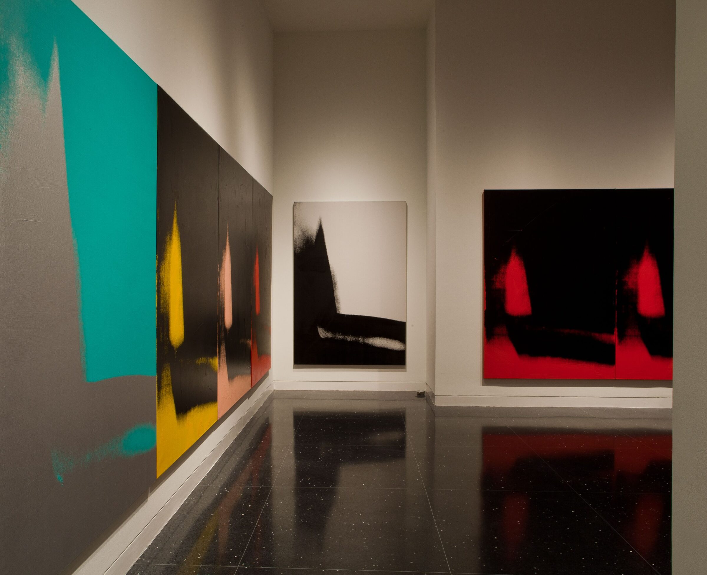 Several mostly black paintings of an identical abstract image in varying colors hang on three white gallery walls.