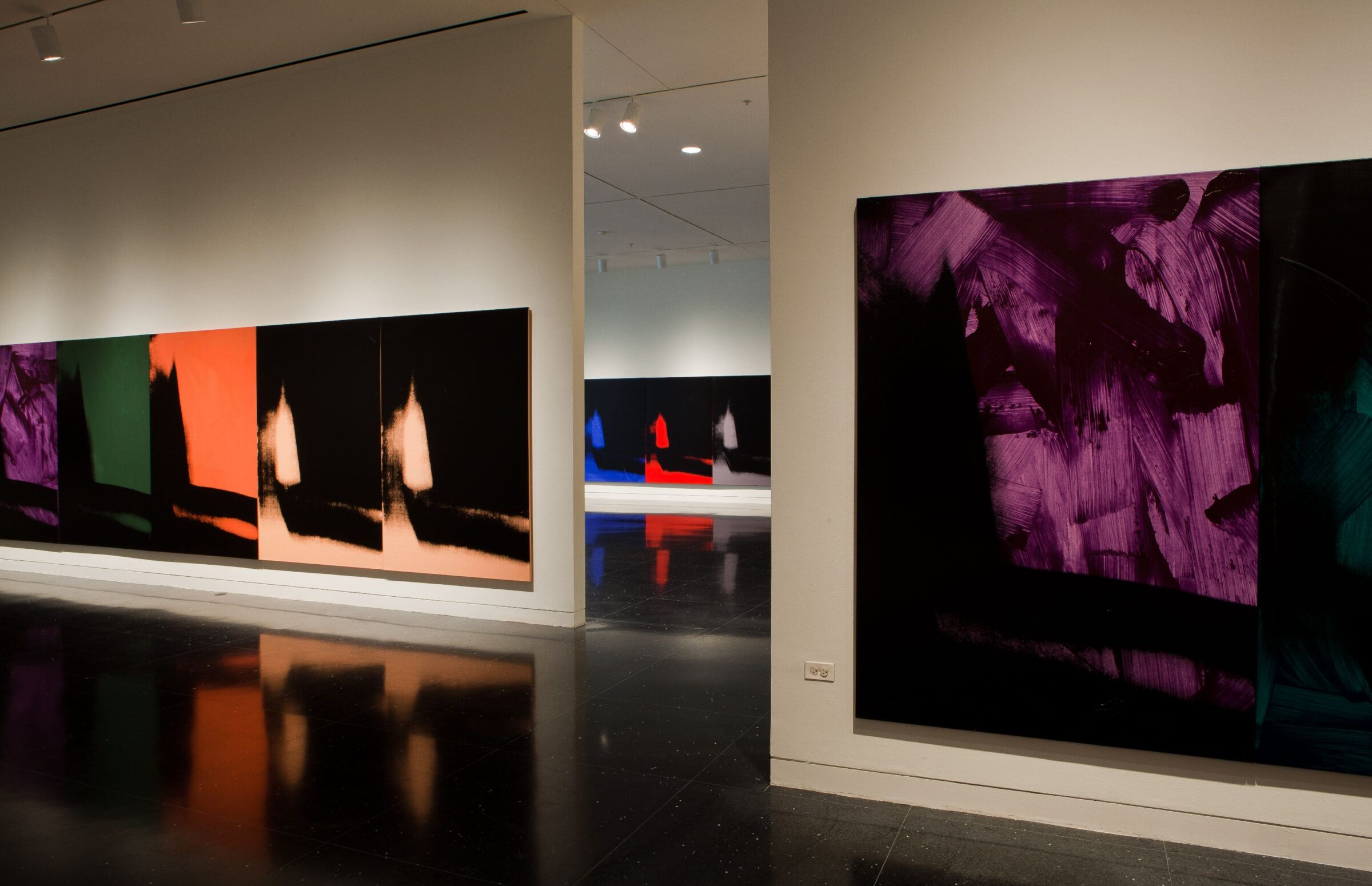 Several mostly black paintings of an identical abstract image in varying colors hang on three white gallery walls.