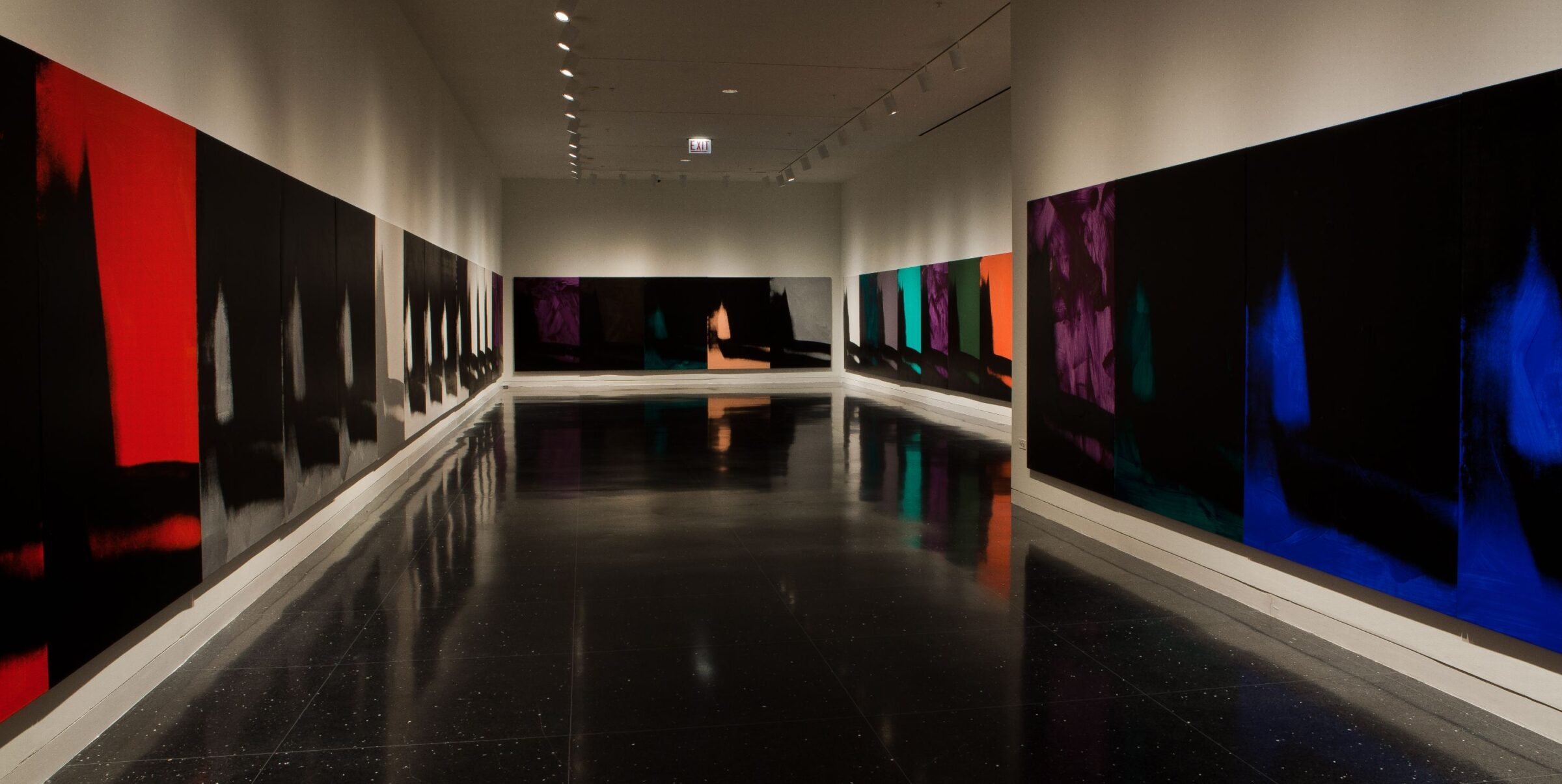 Several mostly black paintings of an identical abstract image in varying colors span the entire length of four white gallery walls.