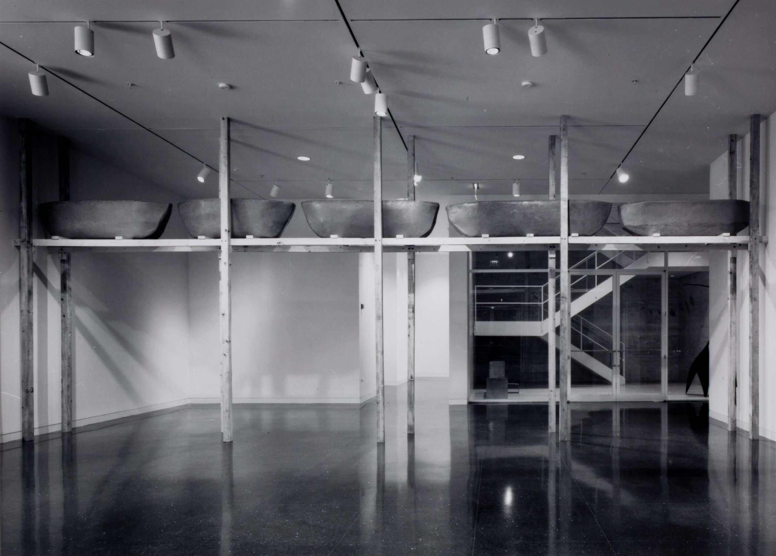 A white gallery space in which five carved boats are installed on a two-by-four scaffolding and nearly touching the ceiling.